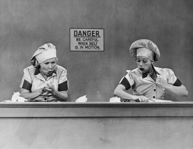Two women in factory uniforms and hats look stressed while working on an assembly line.